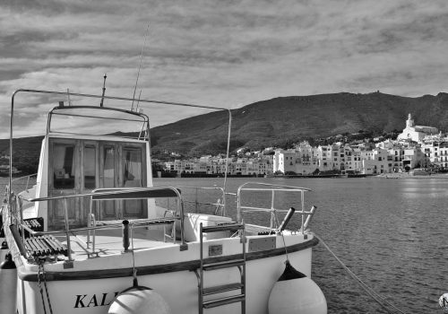 Photographie en noir et blanc d'un bateau amarré à Cadaqués, avec le village en fond.