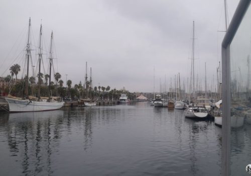 Le port Vell à Barcelone sous le ciel gris avec les mâts des bateaux se reflétant dans l'eau.